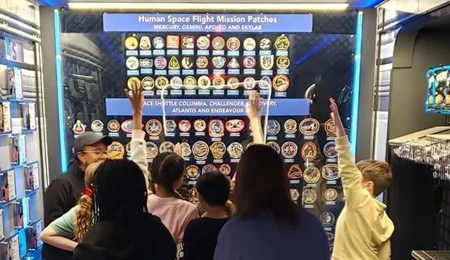 Students, seen from behind, raising their hands at the human space flight mission patches wall inside the trailer